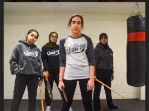 A group of young women of varying races pose with various arnis tools - sticks and daggers - while wearing Sister Fit shirts. 