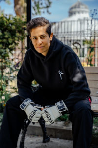 A woman in a black zip up hoodie and gloves used in martial arts sits with an open posture looking directly into the camera.